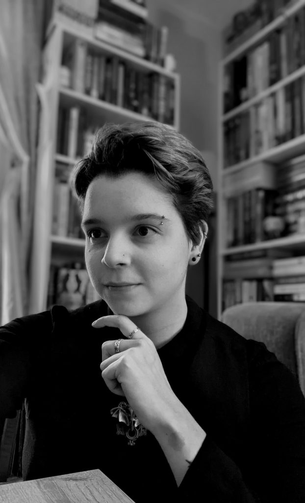 A black and white photo of Talli posing in front of their bookshelves, with a black shirt on, raising a hand to their chin for the pose.