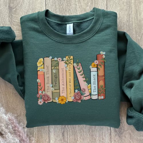 A neatly folded forest green sweater laying over a light wooden background. The sweater is printed with the design of a flowery bookshelf and includes several classic book titles.