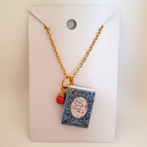 A white paper holds a golden link necklace, and two charms hang from the necklace. The smaller charm on the left is a red and gold sphere, while the right charm is shaped like a blue book cover and reads, "The Secret Garden".
