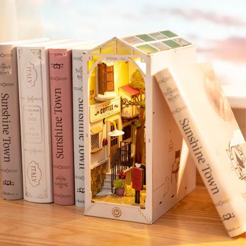 A three-dimensional bookshelf nook stands between several book spines on a wooden surface. The inside of the book nook shows a miniature scene of a European street, which includes a paved stone path, windows, and warm light.