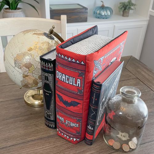 A hyperrealistic novelty pillow in the appearance of a copy of Dracula by Bram Stoker. The pillow stands between two genuine copies of the book as well as a globe and jar of coins on top of a wooden table.