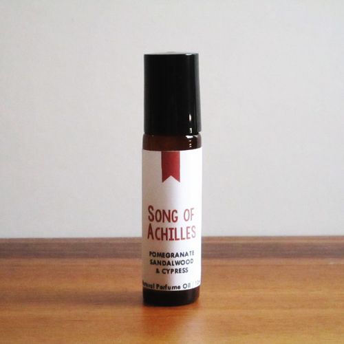 An amber vial of perfume oil with a black cap standing on a wooden table. A white label on the vial reads, in red text, "Song of Achilles", followed by black text that reads, "Pomegranate, sandalwood, & cypress".