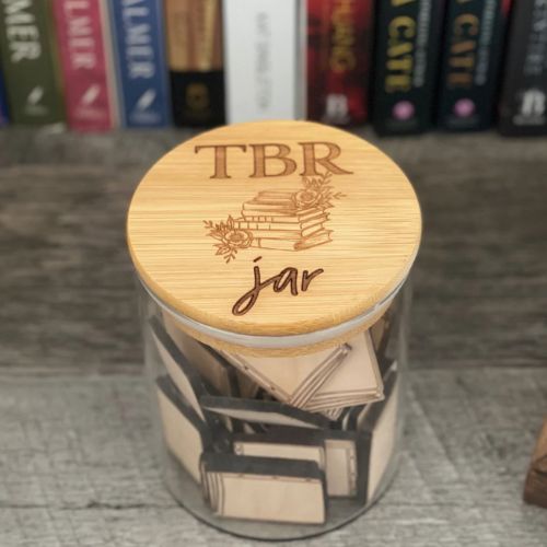 A jar of wooden book-shaped tokens covered with a lid that is decorated with an image of books and flowers and reads, "TBR Jar". The jar stands on a wooden surface, and book spines are lined up behind it.