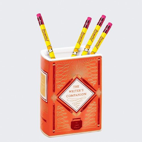 A red vase in the shape of a book is accented with gold and white patterns and reads, in gold text, "The Writer's Companion". The text is surrounded by an illustrated diamond made of ink pens and a pot. In the vase are four yellow pencils.