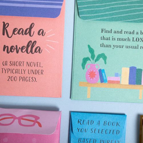A close up of multicolored craft paper pockets on a blue surface. The top left pocket, which is pink and purple, reads in black text, "Read a novella (a short novel, typically under 200 pages)". The top right pocket is green and features an illustration of a table holding many books.