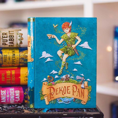 A light blue, rectangular metal tin stands on the edge of a table. The tin is illustrated to resemble a book centering the image of Peter Pan, a young white boy with red hair and leaf clothes. Below is the illustration of a treasure map that reads, "Pekoe Pan". Other tin spines are visible in the background.