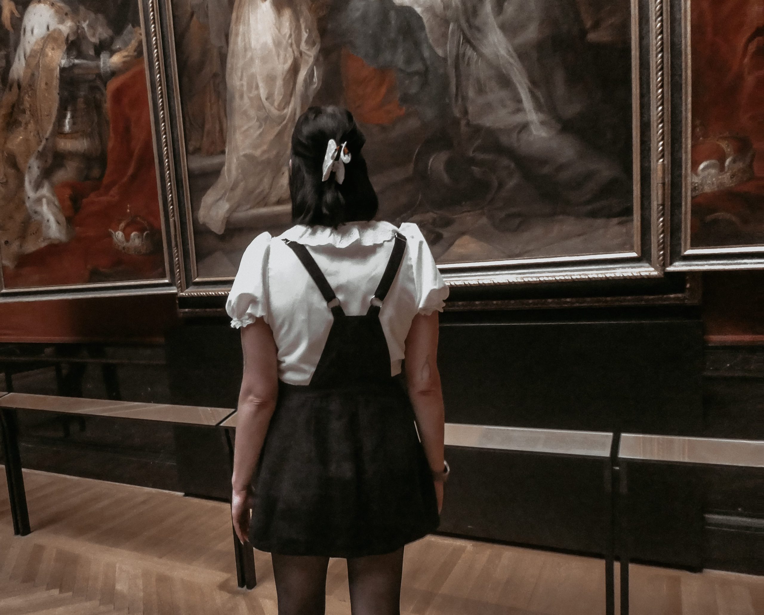Vicky looking at a large, gold framed portrait in a museum with her hair pulled into a half up half down style and a ruffled collared white shirt.