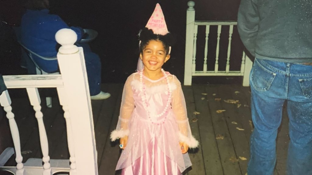 A child Abigail smiling in her pink "Party Princess" costume on a porch with white railings and two adults close by.