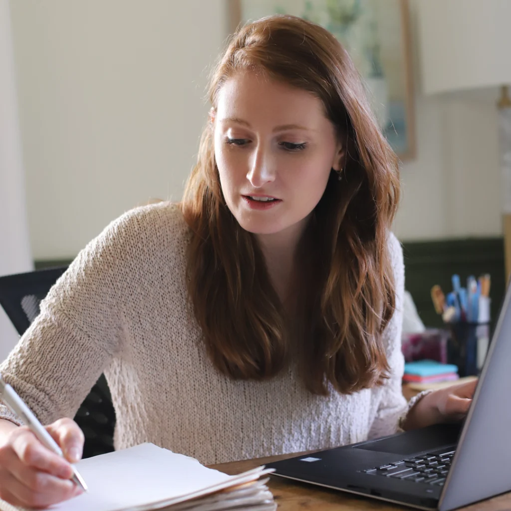 Delemhach writing in what looks to be her office, styling a white shirt and her hair down.