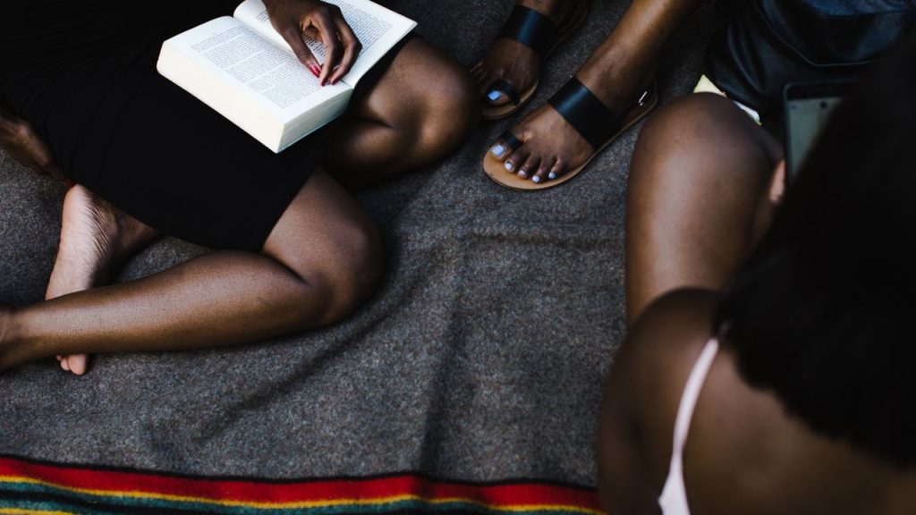 Three people read a book on a black rug with red, yellow, and green stitching