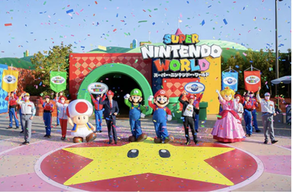 Red and yellow blocks surround a green tube tunnel. The characters Mario, Luigi, Toad, and Peach stand in front of it with cast members around them and colorful confetti.