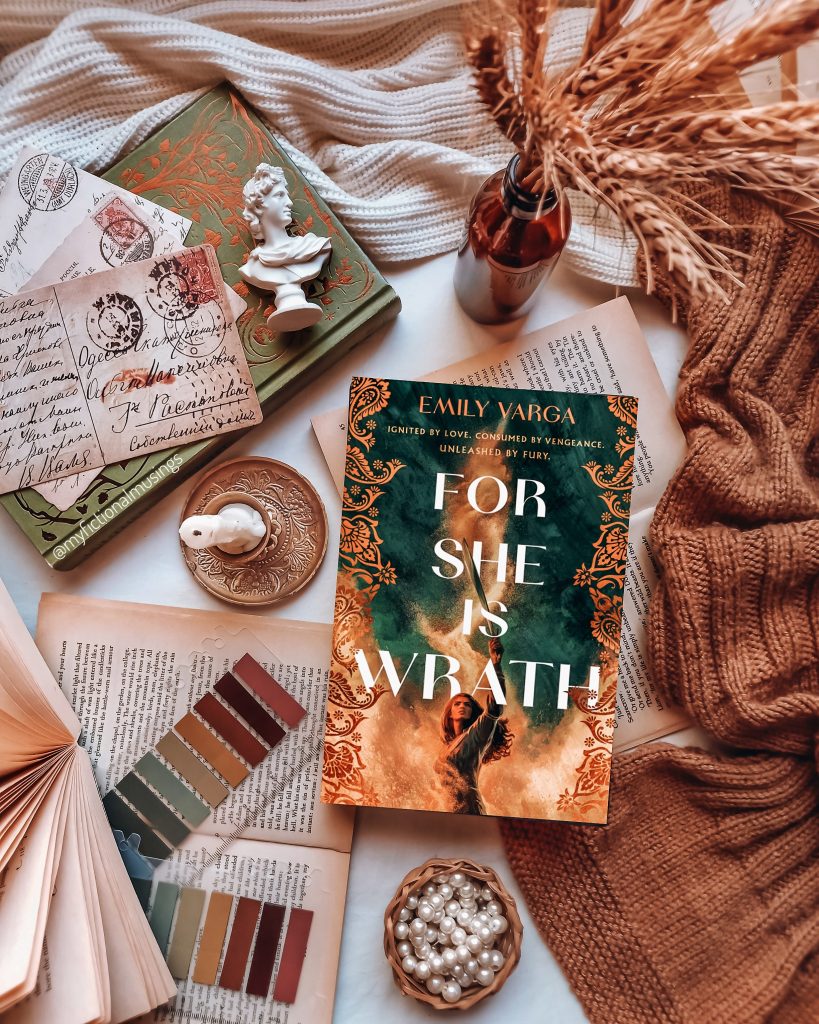 The book "For She Is Wrath" sits on top of other opened books and blankets with incense sticks, postcards, and a candle nearby.