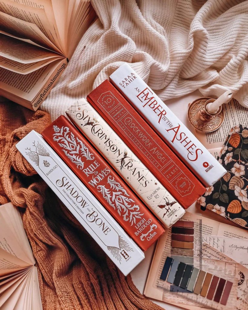 A stack of red and white fantasy books is on its side on a bed. Fanned open books, book markers, and a candle surround it.