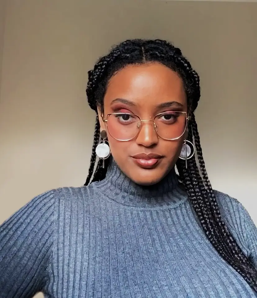 A headshot of a young black woman with long braided black hair and dark brown eyes who is raising an eyebrow. She wears gold glasses, a blue-grey turtleneck sweater, and dangly circular silver earrings. Behind her is a beige wall.