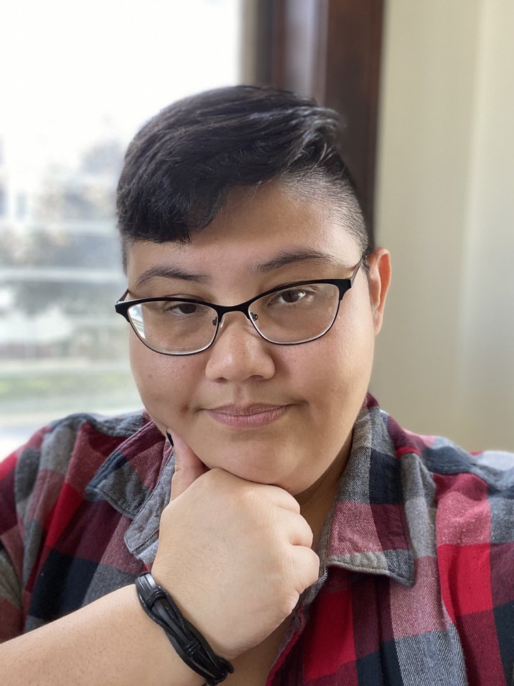 A headshot of a plus-sized Filipino man with cropped black hair and brown eyes. He wears black glasses, a red and grey flannel button down, and a black leather bracelet. Behind him is an open window.