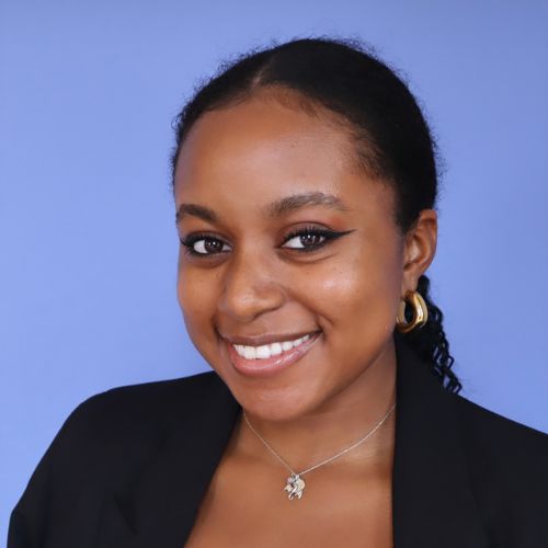 A headshot of a young Black woman with black hair and brown eyes who is smiling. She wears a white corset top, a black blazer, a silver dragon necklace, and small gold hoop earrings. Behind her is a blue background.