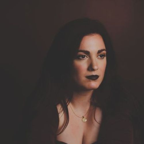 A headshot of a young white woman with long brown hair and dark eyes who is looking off to the side. She wears dark lipstick, a long-sleeved brown top, and a gold necklace. Behind her is a brown background.