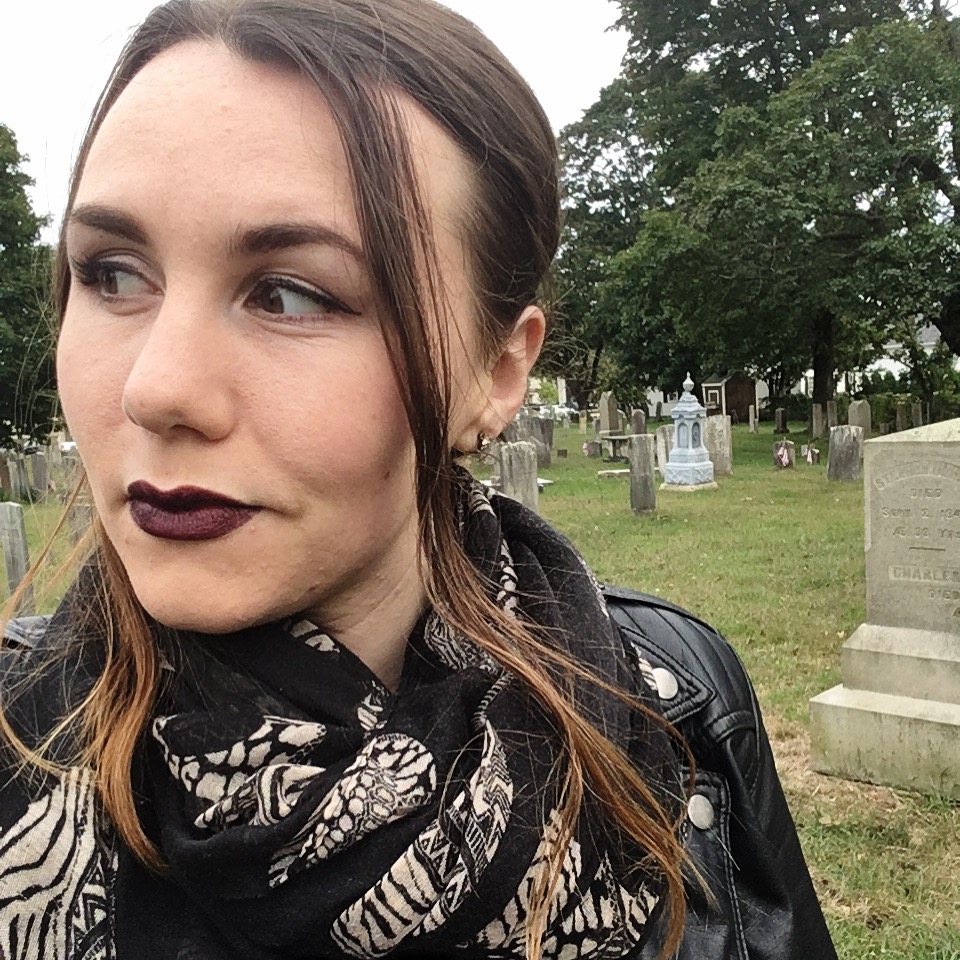 A headshot of a young white woman with long brown hair and brown eyes who is looking off to the side. She wears dark lipstick, a black and white patterned scarf, and black leather jacket, and small silver earrings. Behind her is a graveyard.