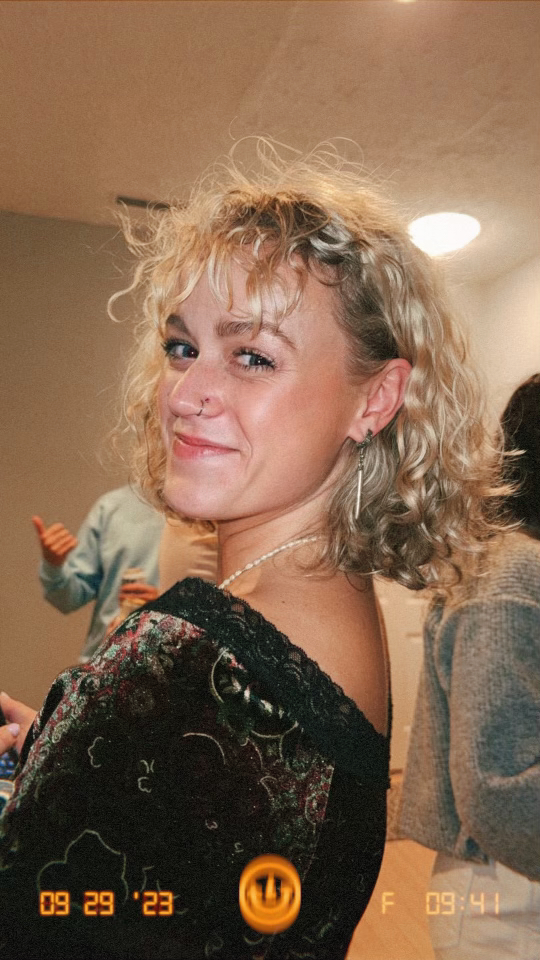 Orianna McDavid giving an over-the-shoulder smirk; she's in a beige room surrounded by a few people.