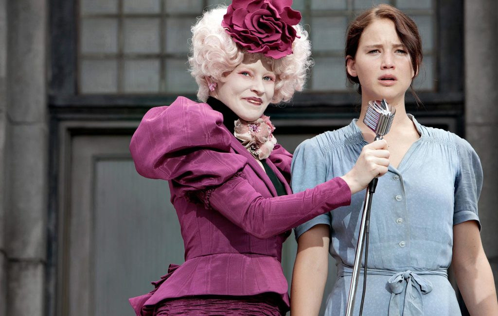 A woman in extravagant pinkish purple costume holds a mic to a plainly dressed woman.
