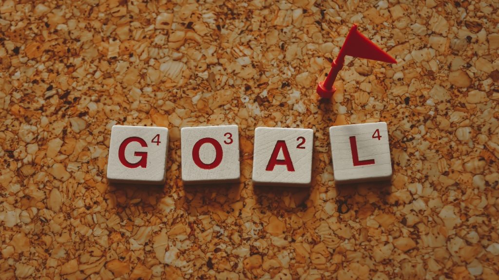 Goal is written out using wooden scrabble tiles next to a tiny plastic red flag all on a corkboard background.