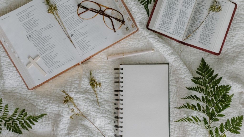 A notebook, and 2 other books sitting on a blanket with a pen glasses and various leaves