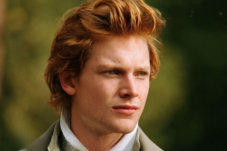 A young white man with ginger hair and freckles. He is looking off to the side and appears to be standing near greenery.