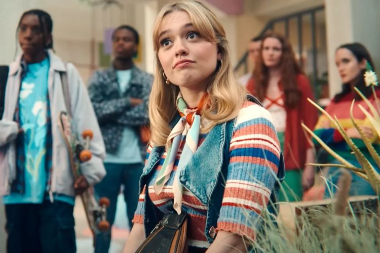 A young white woman with blonde hair and colorful striped clothing. She is looking in surprise up at something off to the side, and there are other people standing behind her.