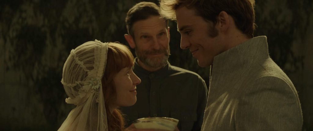 Finnick and Annie standing before a minister during their wedding ceremony with Annie in a white vail and dress and Finnick wearing a grey suit with the minister wearing a dark grey suit.