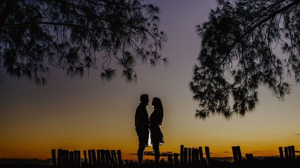 A man and a woman are standing close to one another, gazing into each other's eyes. There is a sunset and a view behind them.