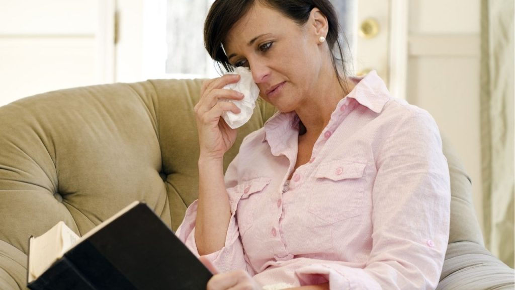 A woman in a pink shirt is tabbing a tissue to her eyes, tearing up while reading a book on a couch.