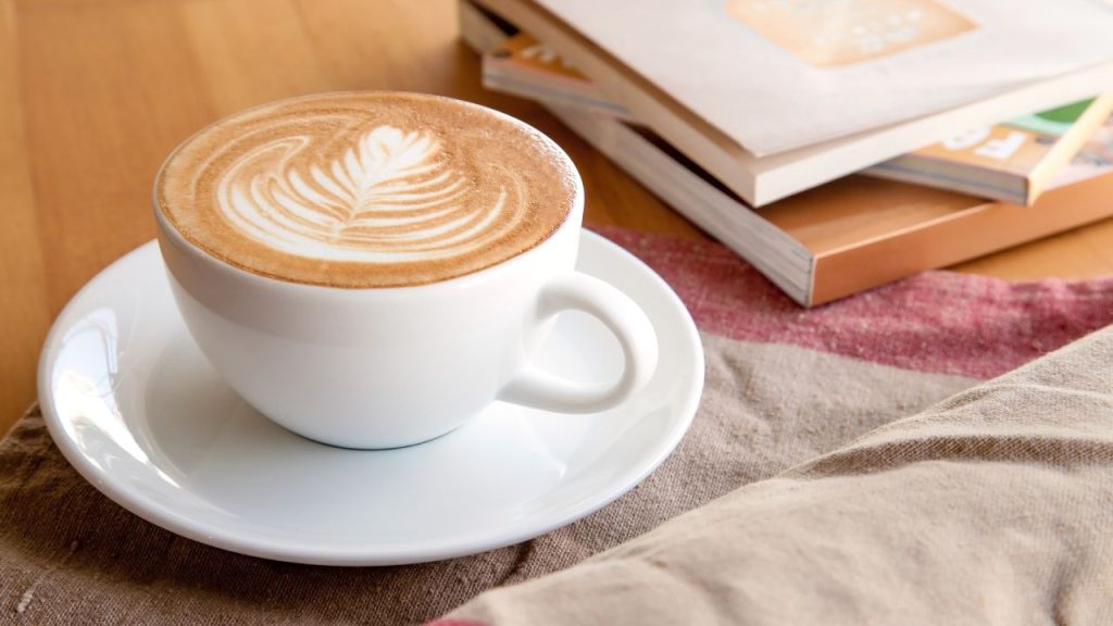 A coffee with a flower design on top with a stack of books and a table cloth on a table.