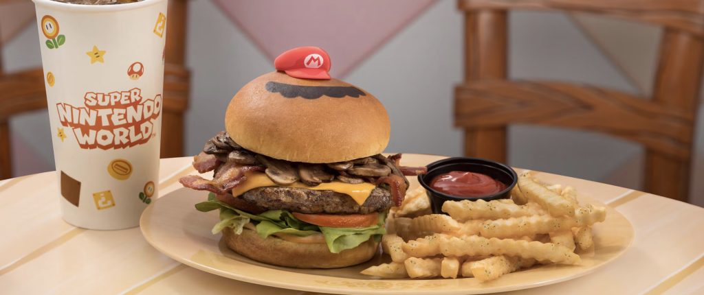 A mushroom burger with fries and a drink. The burger has a plastic red Mario hat and his black mustache on top of it.