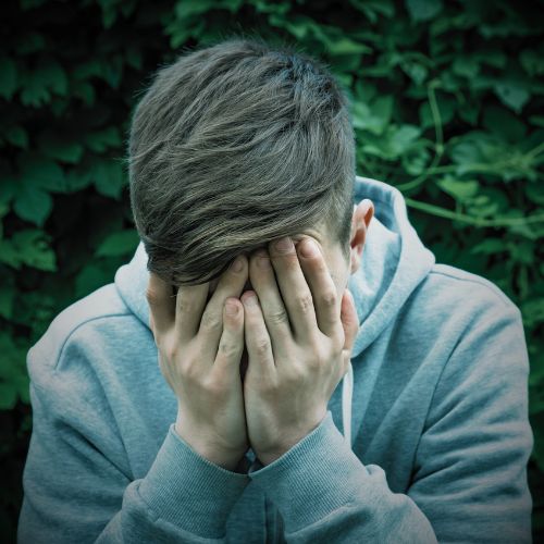 A masc-presenting teen wearing a gray hoodie sits outside with their hands covering their face in distress.
