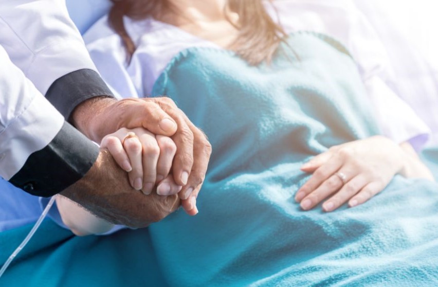 Someone lying in a hospital bed with one of their hands clasped by someone else's.