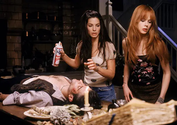 Two women standing over a man laying on a table and a single candle besides him. One woman holding a can of whipped cream