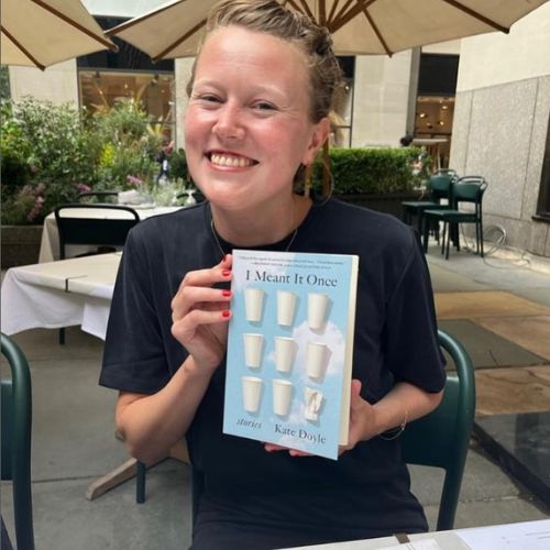 Kate sitting at an outside bistro table holding her book smiling at the camera