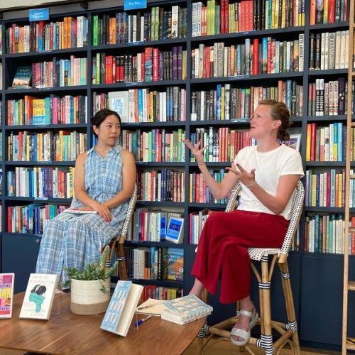 Kate sitting in front of a large book case alongside an asian woman animatedly talking.