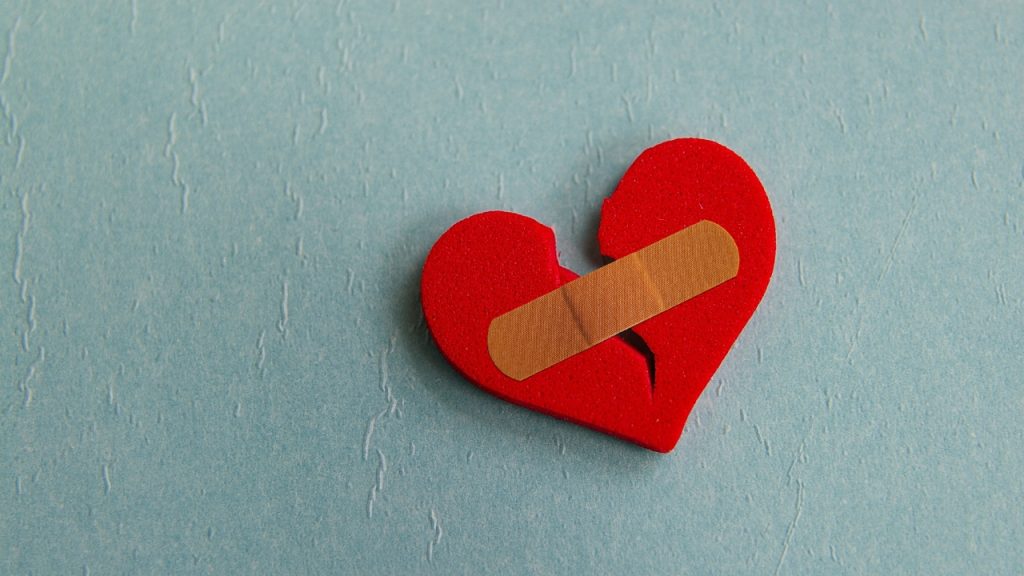 Broken heart with a bandage over it on a seafoam blue background.