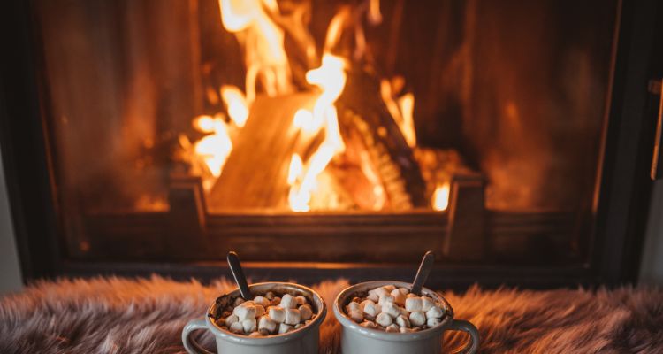 A fire in the fireplace burns bright as two hot cocoas with marshmallows sit on a fuzzy blanket.