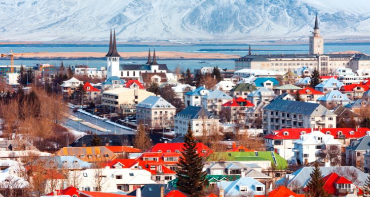 A wide view of a town is shown. Many of the buildings have red roofs. There is a body of water and a mountain in the distance.
