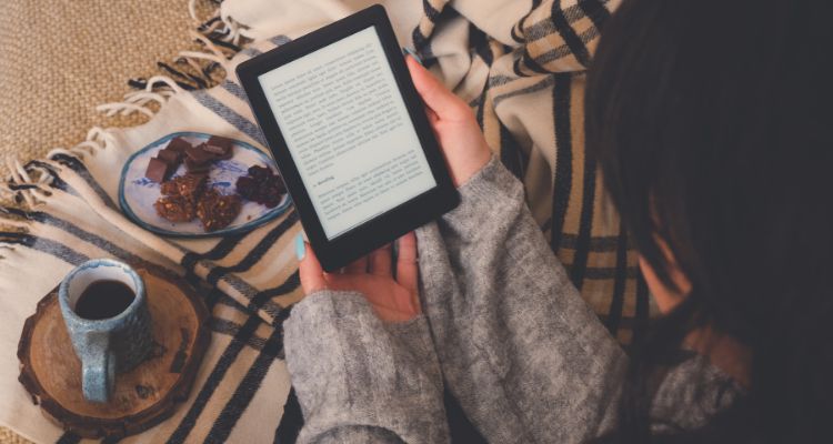 A woman in a gray sweater stares down at her black-trimmed ebook. She is on a beige blanket with a cup of coffee and some chocolate.