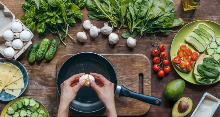 A person cracks an egg above a pan. Around them are various veggies and ingredients for their cooking.