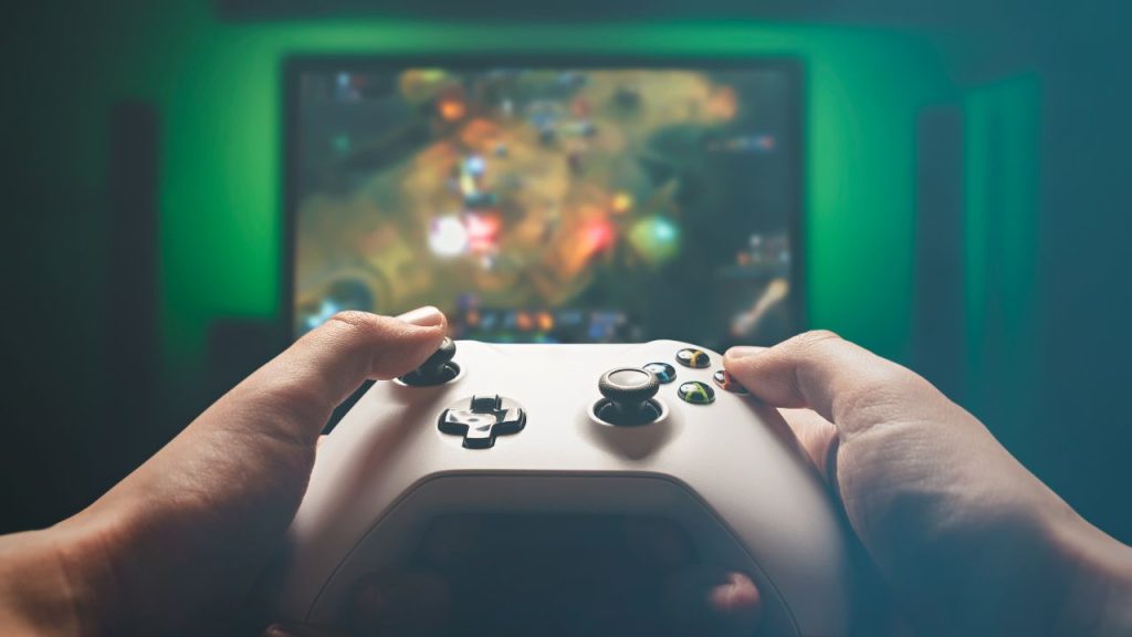 Two hands hold a white Xbox control in front of a TV screen with green lighting as they play a video game.