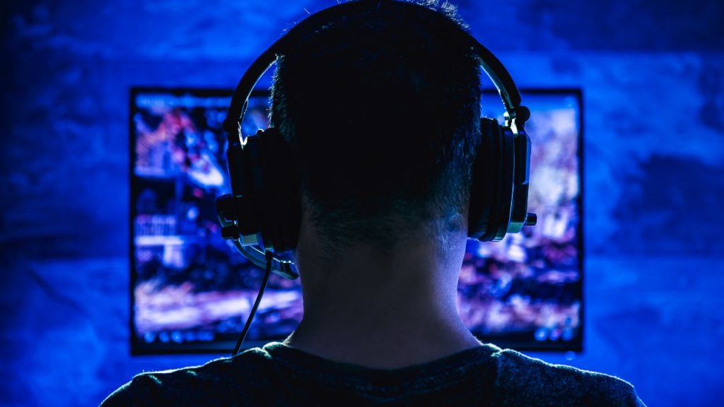 A figure sits with their back to the camera as purple light emanates from their screen as they play an online game.