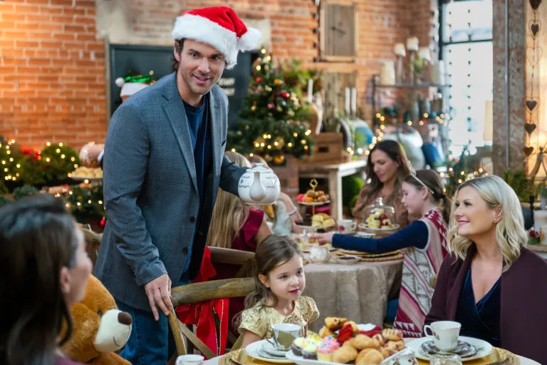 Scene from 'Checkin' it Twice.' Scott holding a teapot talking to a table with Ashley, another woman, and a little girl.