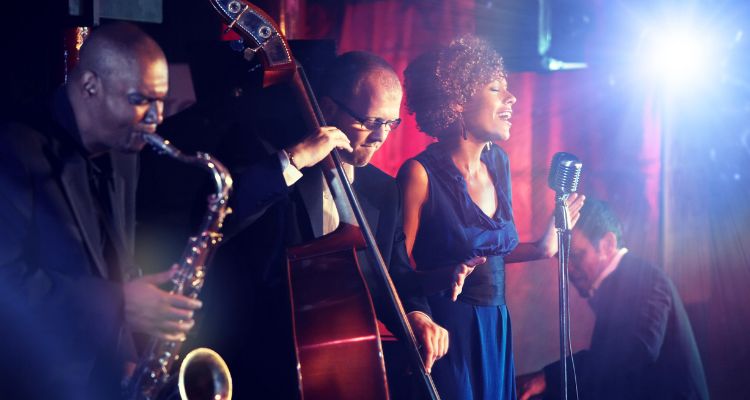 A group of male jazz musicians in suits play saxophone, upright bass, and piano as a woman in a blue dress sings into an old-fashioned microphone.