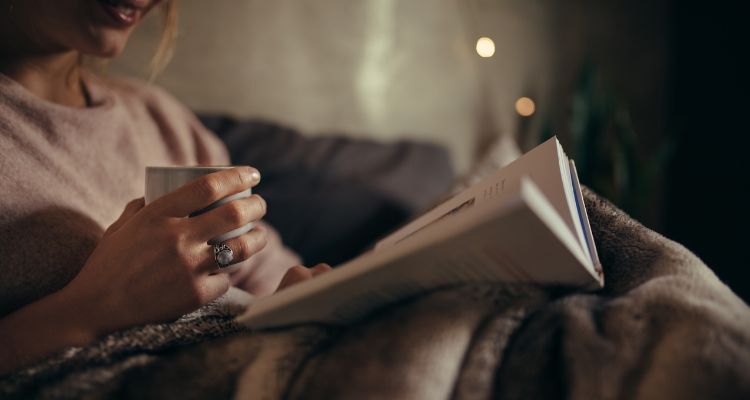 A person smiles and reclines under the covers in bed while holding a stoneware cup in one head and holding a book open on the blankets in the other.