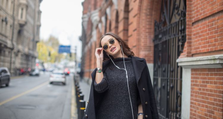 A woman in black coat over a sweater and wearing wired white earbuds struts down a city street and smiles while adjusting her sunglasses with one hand.