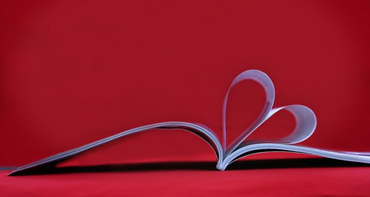 Pages of an open book have been curved inward and tucked into the spine to form the shape of a heart when viewed from beneath, against a red background.